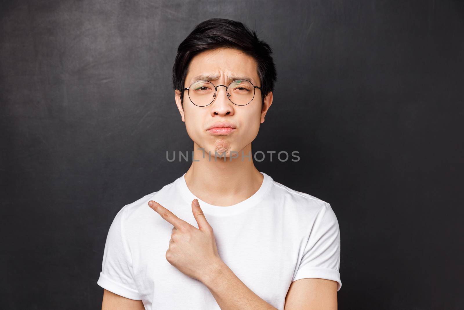 Close-up portrait of picky, skeptical and gloomy asian guy in glasses, grimacing unsatisfied, look disappointed at camera while pointing finger left at something unpleasant and uncool.