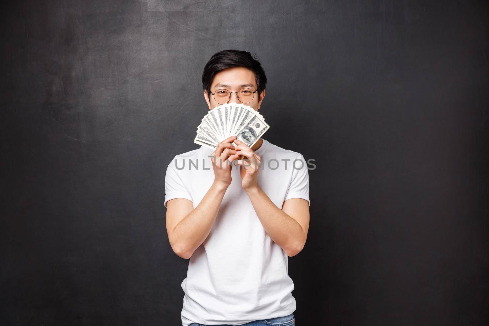 Portrait of sly happy and pleased asian young man winning big cash prize money, hiding face behind fan of dollars smiling with eyes, decide what to buy on it, standing black background.