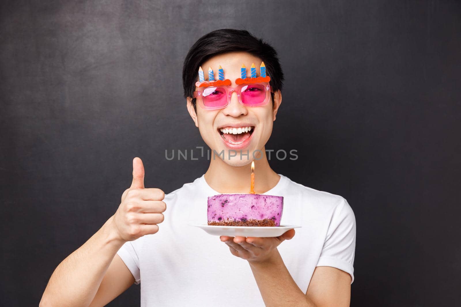 Celebration, holiday and birthday concept. Close-up portrait of excited happy b-day guy in funny party glasses, show thumb-up and hold delicious cake, making wish on lit candle, black background.