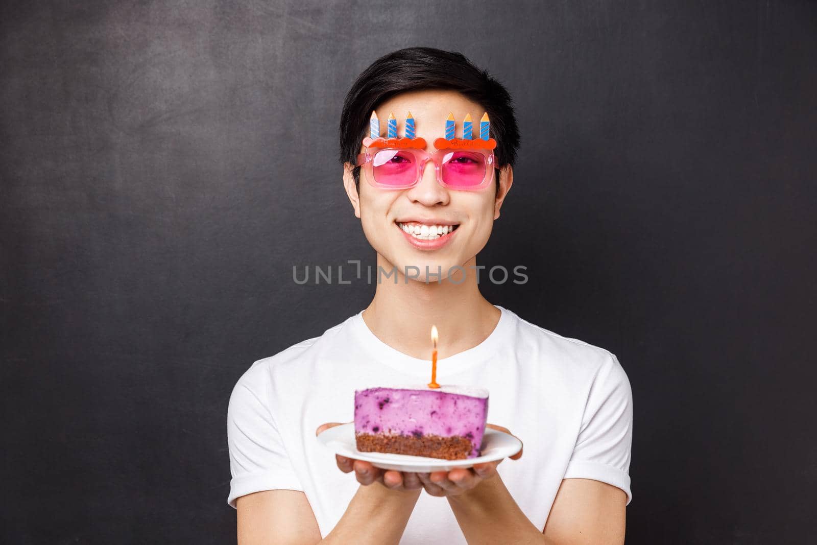 Celebration, holiday and birthday concept. Close-up portrait of excited and happy, cheerful asian man celebrating b-day in funny glasses, holding cake, blow-out candle, black background.