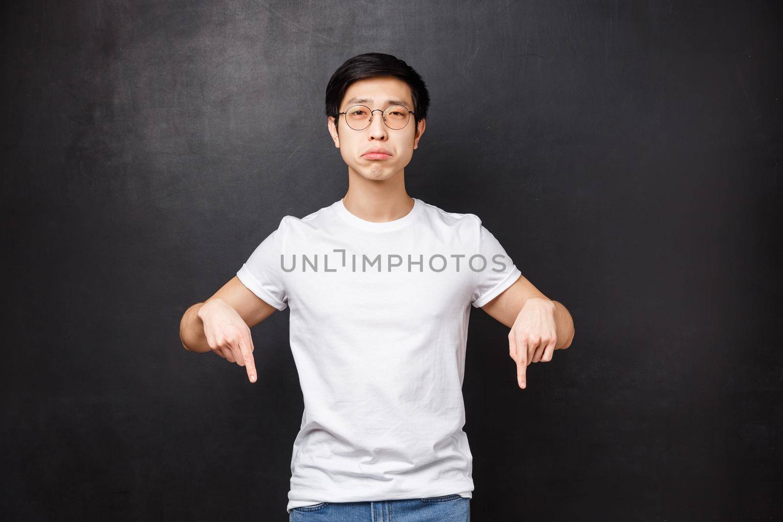 Miserable and gloomy upset cute asian guy sobbing look at camera with regret and sadness, pointing fingers down at something broken or upsetting, feel uneasy, black background.