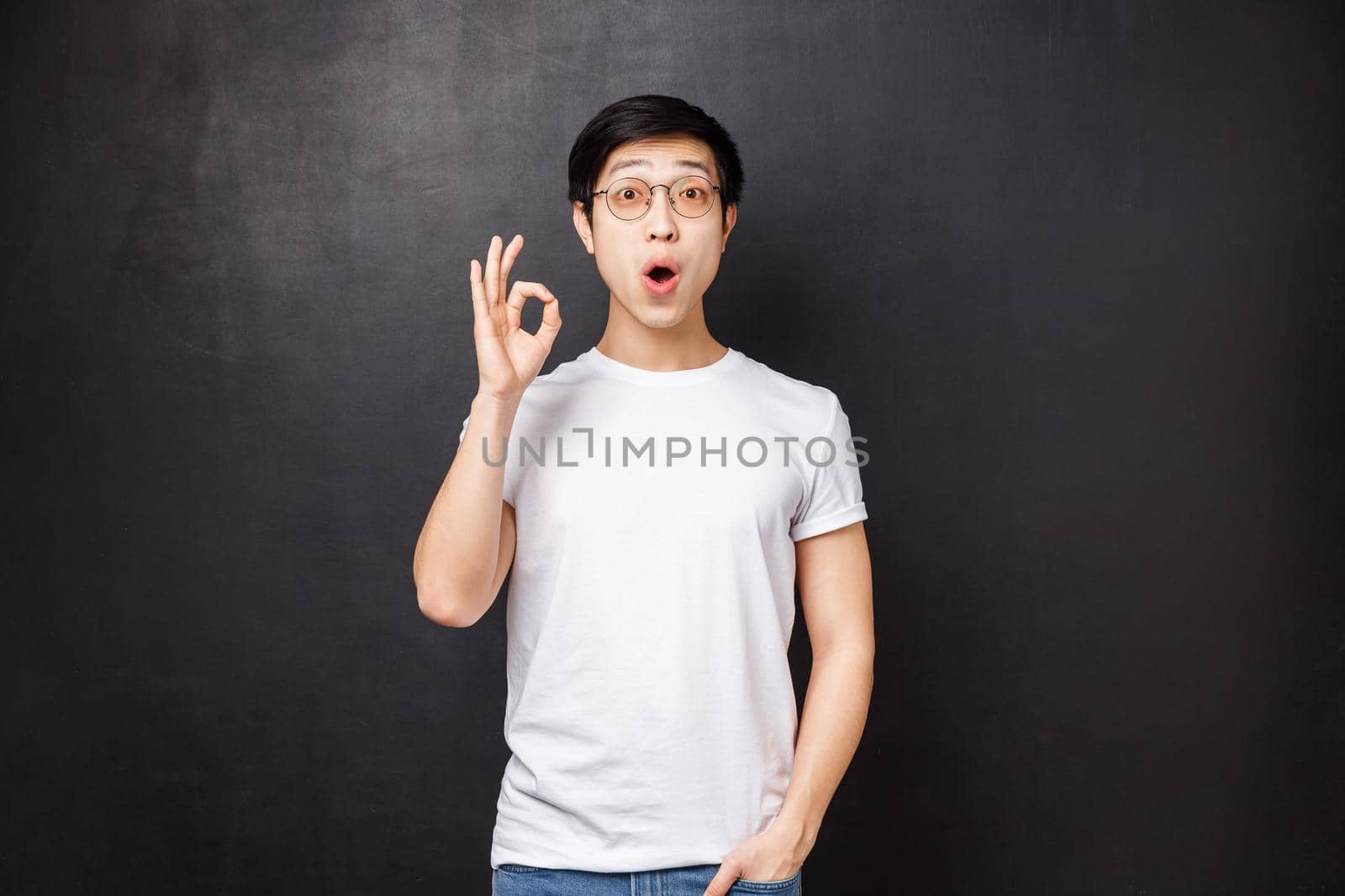 Amazed and enthusiastic young surprised asian guy left speechless and impressed after seeing premier of new movie, show okay sign look astounded standing black background.