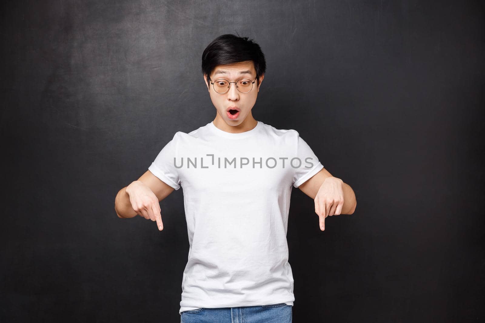 Astonished and amazed speechless asian guy see something shocking and awesome, pointing looking down, folding lips amused say wow, stare impressed at cool product, black background.