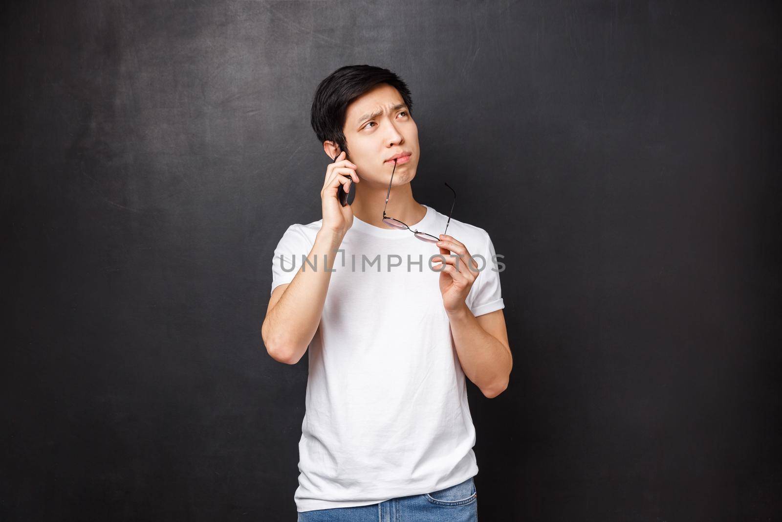 Technology, gadgets and people concept. Portrait of thoughtful and indecisive asian guy biting rim of glasses while thinking as talking on mobile phone, making hard choice what order over smartphone.