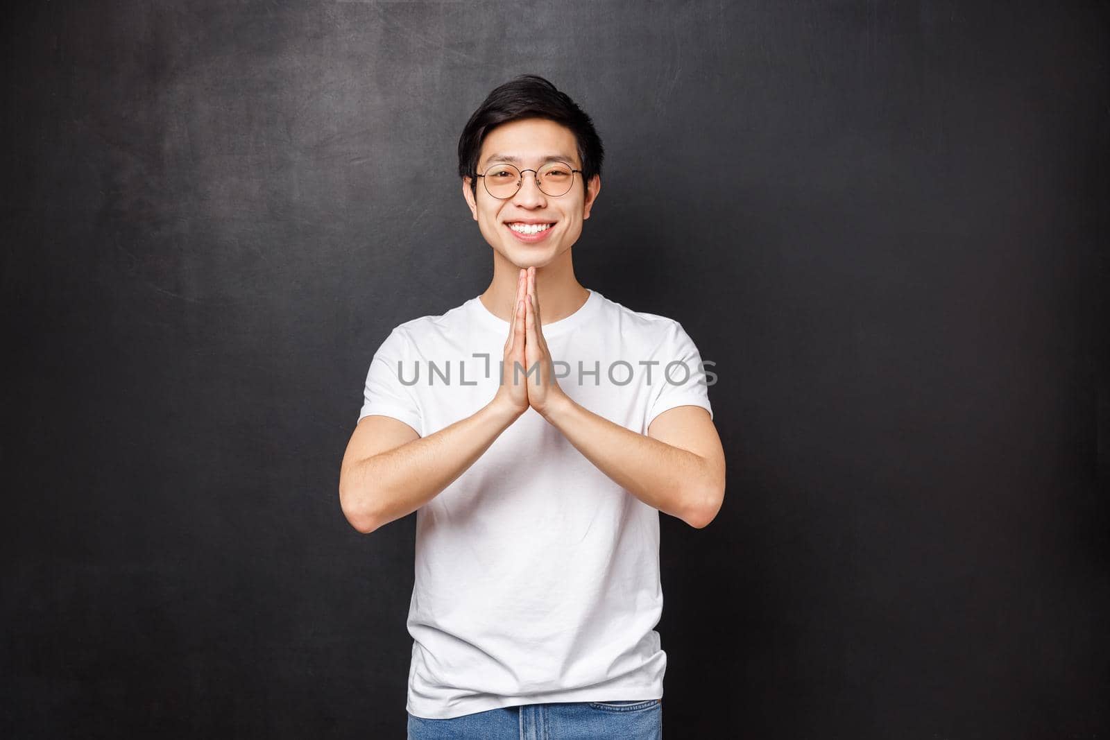 Handsome young asian hipster guy in white t-shirt, glasses, hold hands in namaste, pray gesture smiling grateful, thanking for help, pleading or making wish, standing black background.