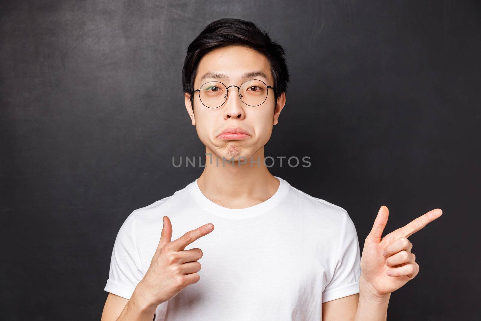Close-up portrait of impressed asian man in glasses white t-shirt, pouting and looking camera with approval, pointing fingers right at really cool awesome product, agree with you, black background.