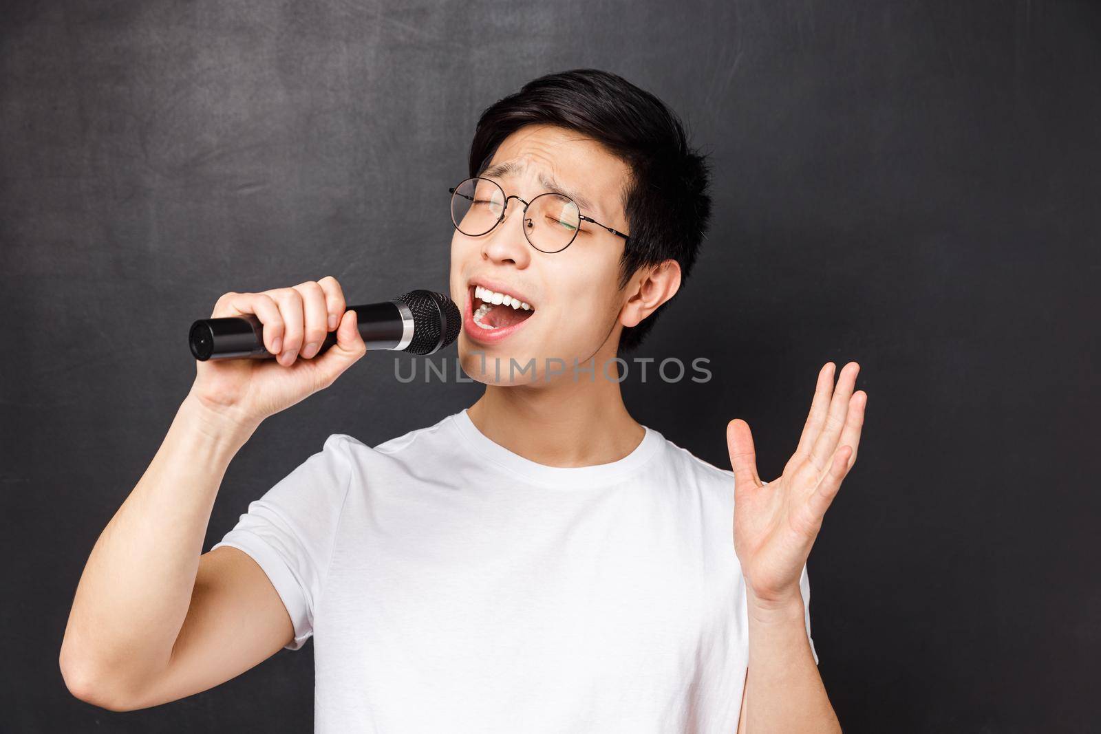 Leisure, people and music concept. Close-up portrait of passionate and carefree asian guy likes singing songs, holding microphone and raising one hand, perform in fron of friends on karaoke party.