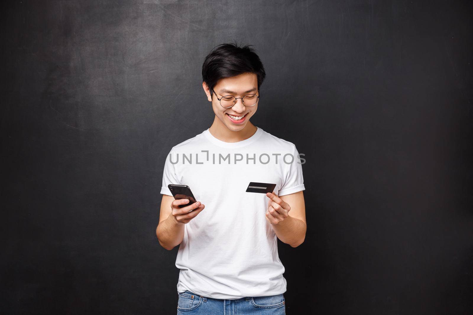 Bank, finance and payment concept. Portrait of cheerful asian man in t-shirt, holding mobile phone and credit card, insert billing info, buying products online, book tickets for flight with app.