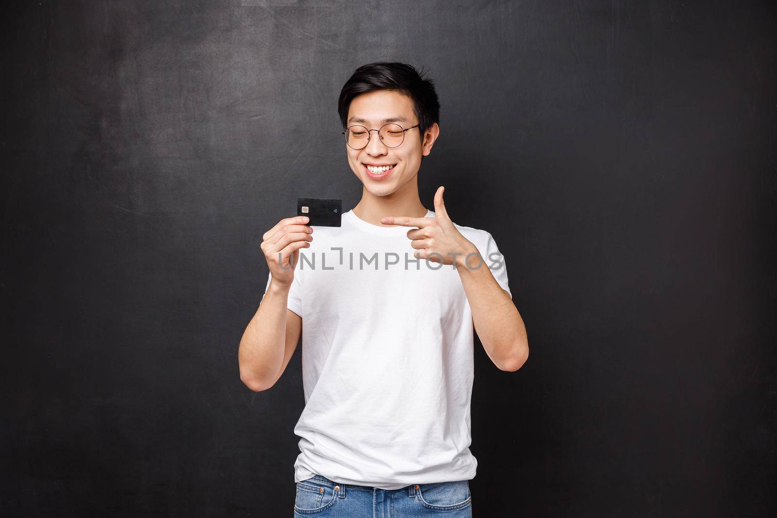 Bank, finance and payment concept. Portrait of good-looking asian male in t-shirt and glasses, talking about credit card features, deposit or cashback, place money on account, black background.