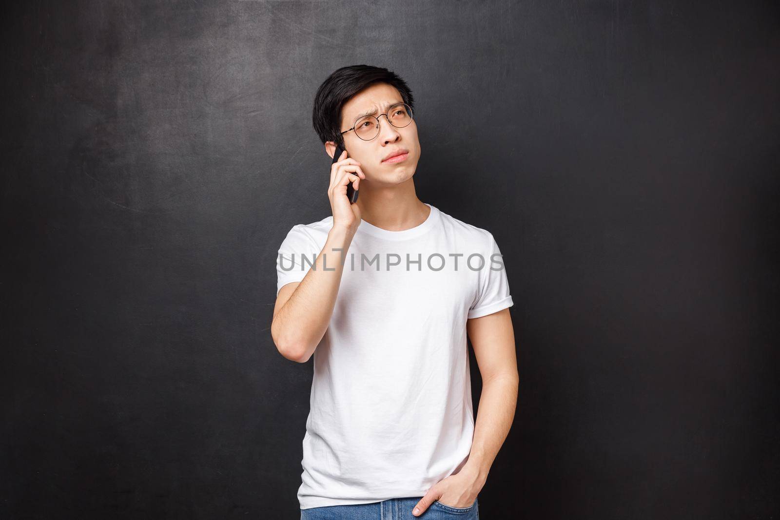 Technology, gadgets and people concept. Portrait of thoughtful young asian guy thinking over question, calling friend, talking on phone, hold smartphone and look away pondering what answer.