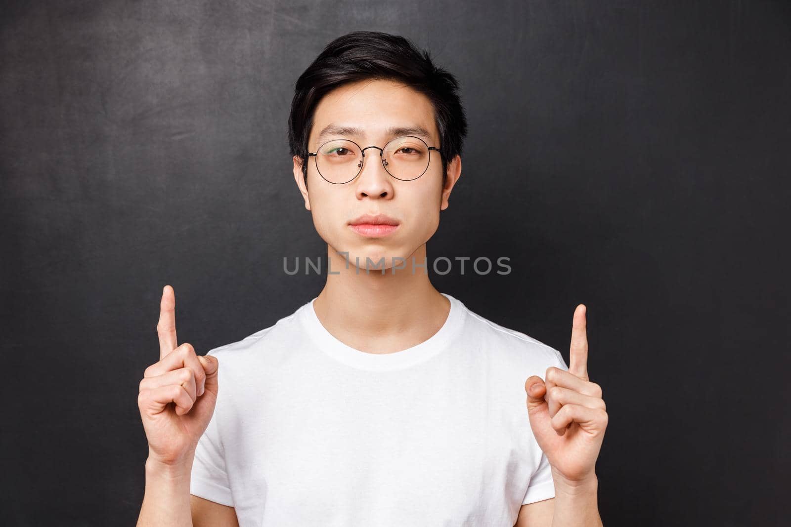 Close-up portrait of serious-looking determined young asian man in glasses and white t-shirt, pointing fingers up and looking straight to camera with no emotions, being focused.