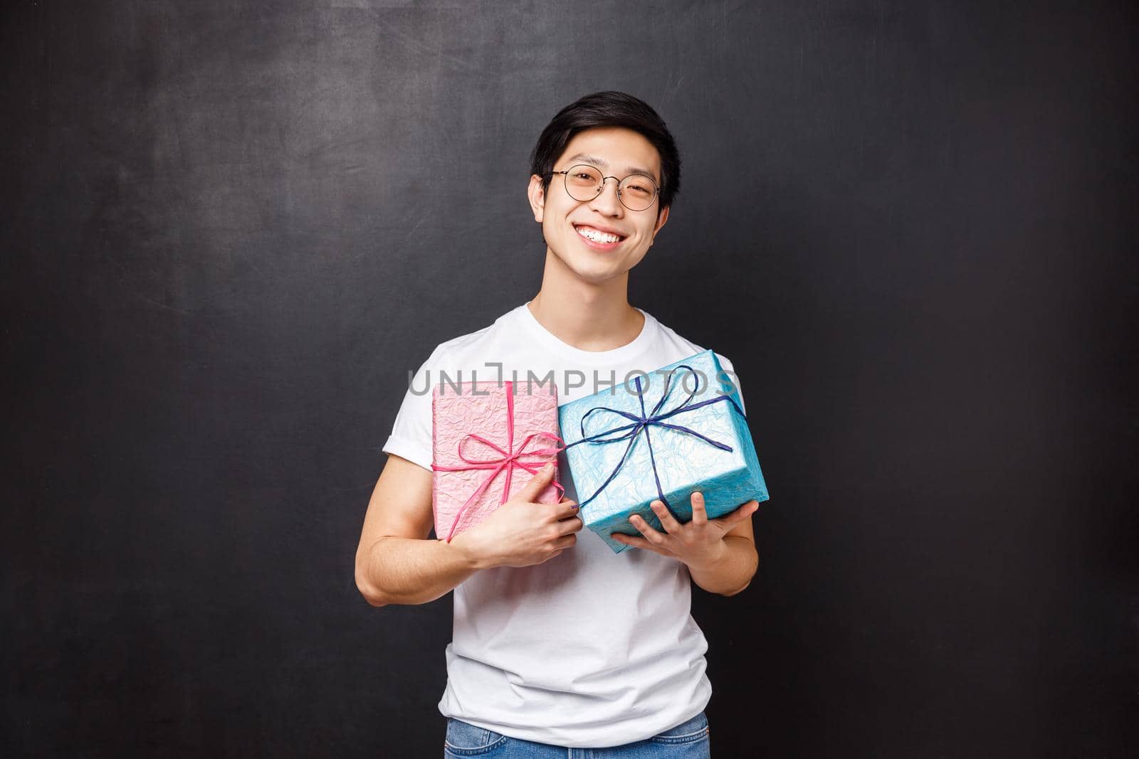 Celebration, holidays and lifestyle concept. Portrait of cute and handsome boyfriend prepared presents for his partner, holding two gift boxes pink and blue, smiling proud, black background.
