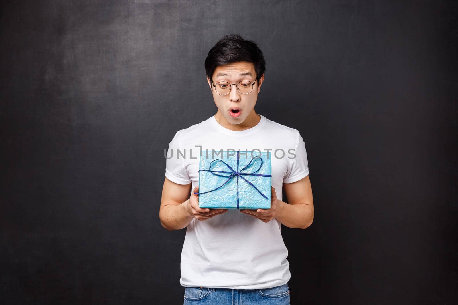 Celebration, holidays and lifestyle concept. Surprised and excited, amazed asian guy receive gift box, holding present and looking at it amused didnt expect coworker remembers about birthday.
