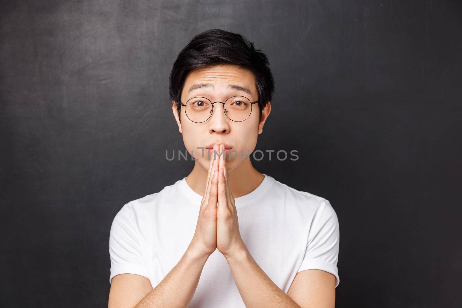 Close-up portrait of worried and uneasy hopeful young man in white t-shirt, glasses, hold hands in pray looking with pleading eyes, asking for help, begging forgiveness, black background.