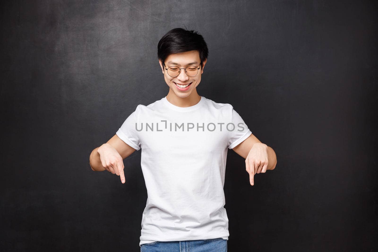 Portrait of cheerful friendly-looking asian hipster guy in sunglasses and white t-shirt spot cool promo discount, got best deal, pointing and looking down satisfied, stand black background.