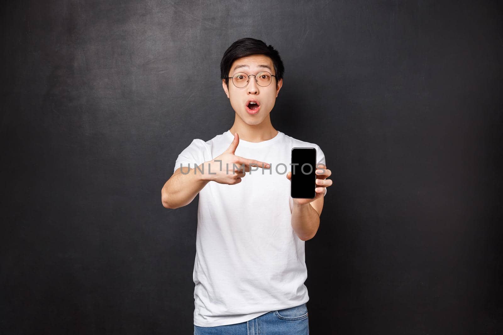 Technology, gadgets and people concept. Impressed and excited handsome asian guy telling to download app, check and see awesome application in your mobile store, point at smartphone screen.