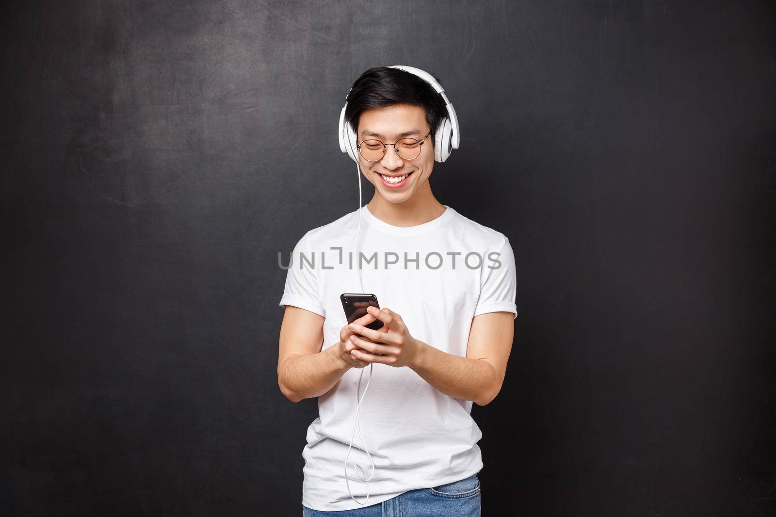 Technology, gadgets and people concept. Handsome happy young smiling asian man in t-shirt, listen music in headphones, pick playlist in mobile phone, texting friend, black background.