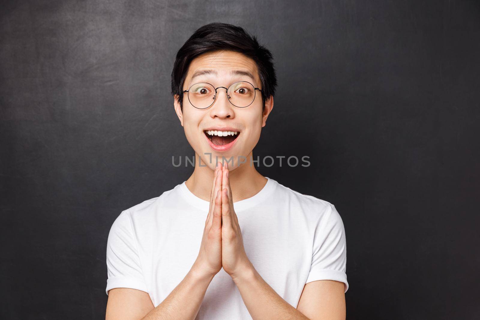 Close-up portrait of hopeful and grateful young happy asian guy receive help after begging friend, saying thank you, hold hands in pray, appreciate gesture, standing black background.