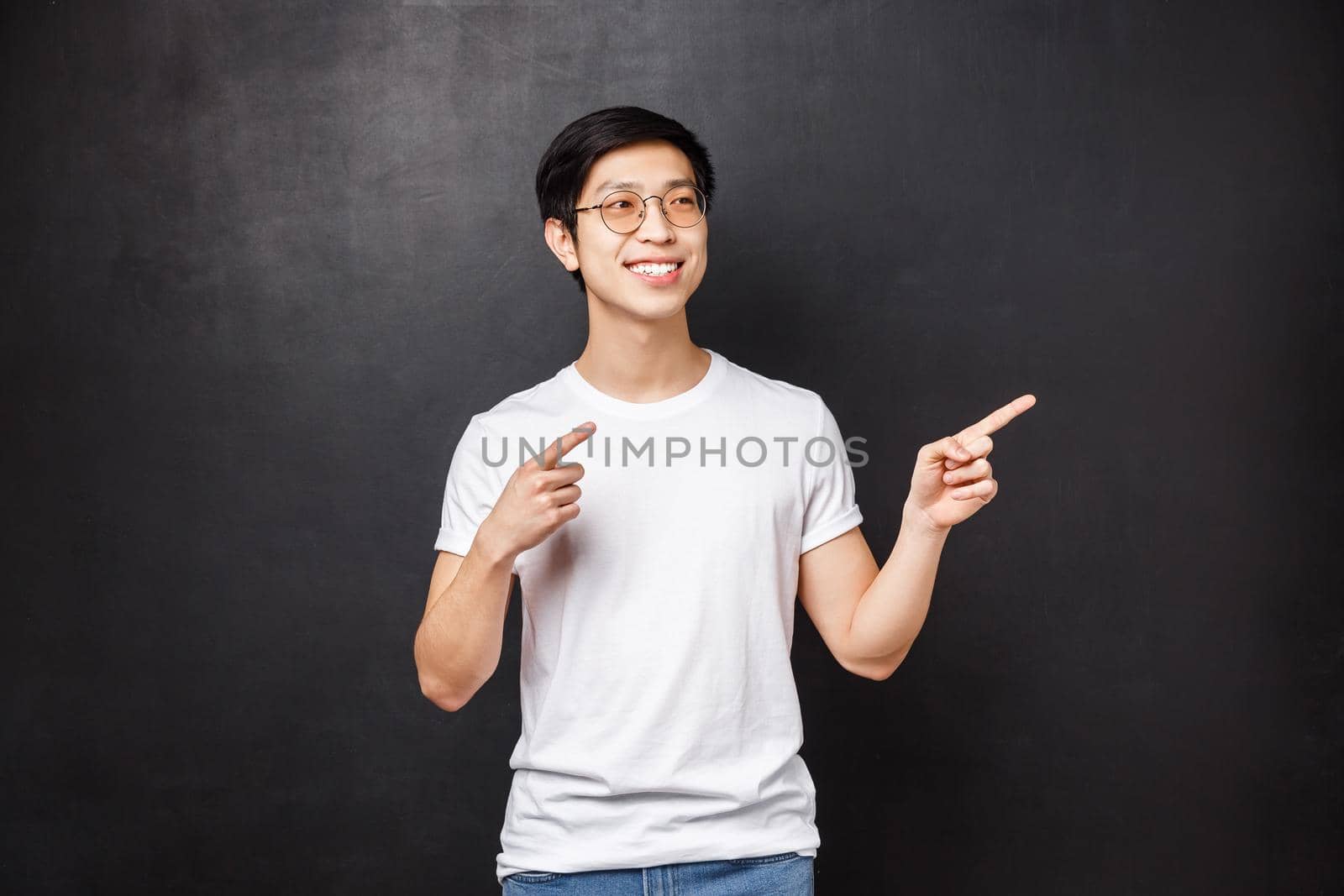 Handsome stylish young asian guy in white shirt, sunglasses pointing looking right side copy space, pleased smile, found exactly what he wanted, choosing this product, black background.