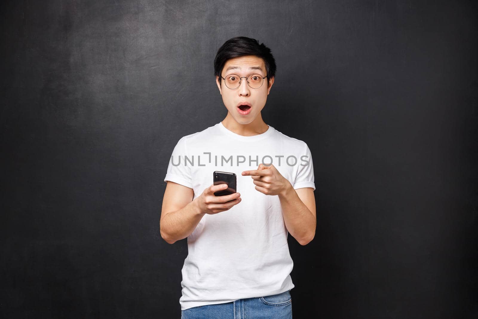 Technology, gadgets and people concept. Impressed and speechless fascinated asian guy saw something super cool in internet, telling about it at camera, open mouth say wow, hold mobile phone.
