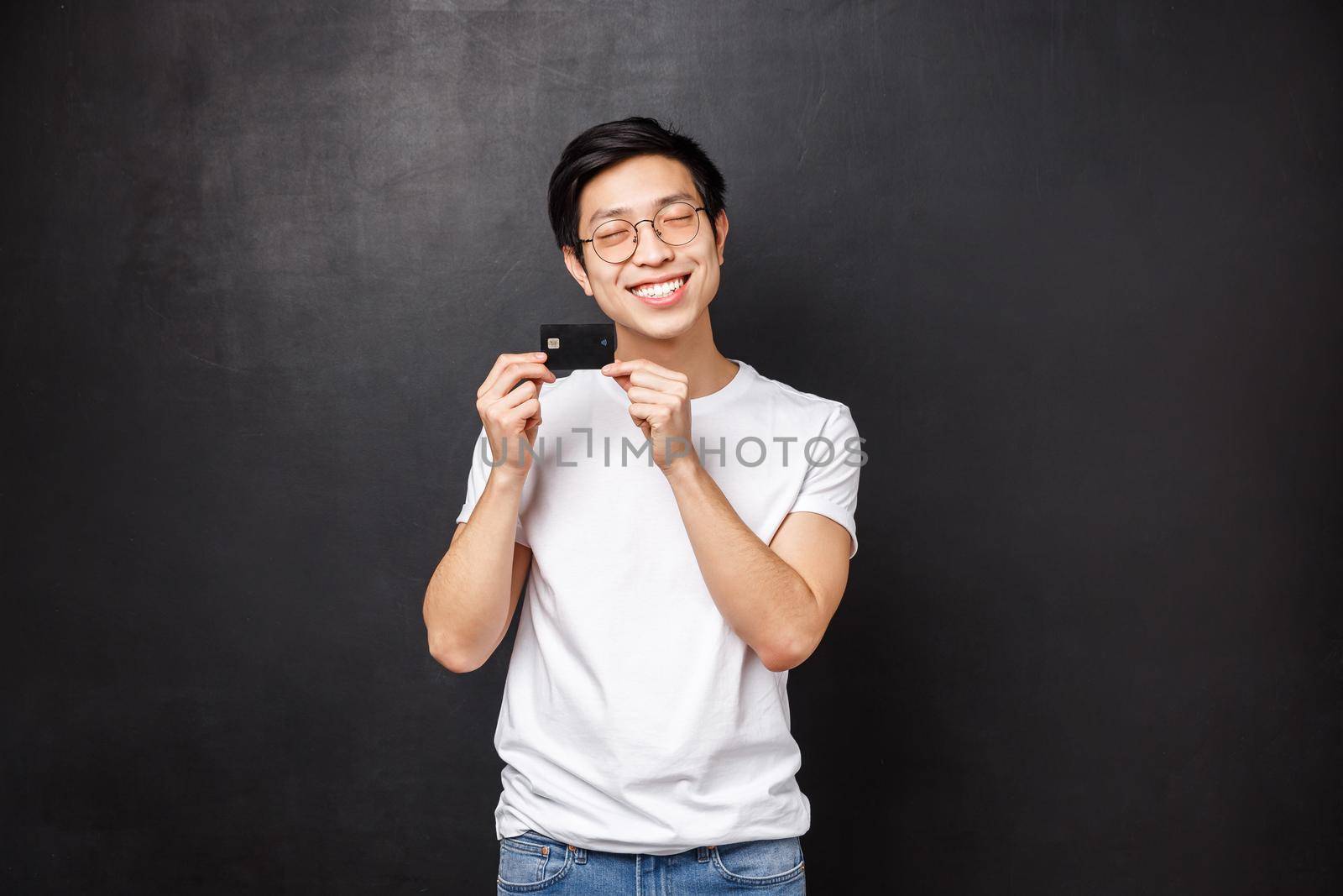 Bank, finance and payment concept. Portrait of silly and happy asian man in t-shirt, close eyes satisfied introducing his first credit card, ready go shopping and waste some money, black background.
