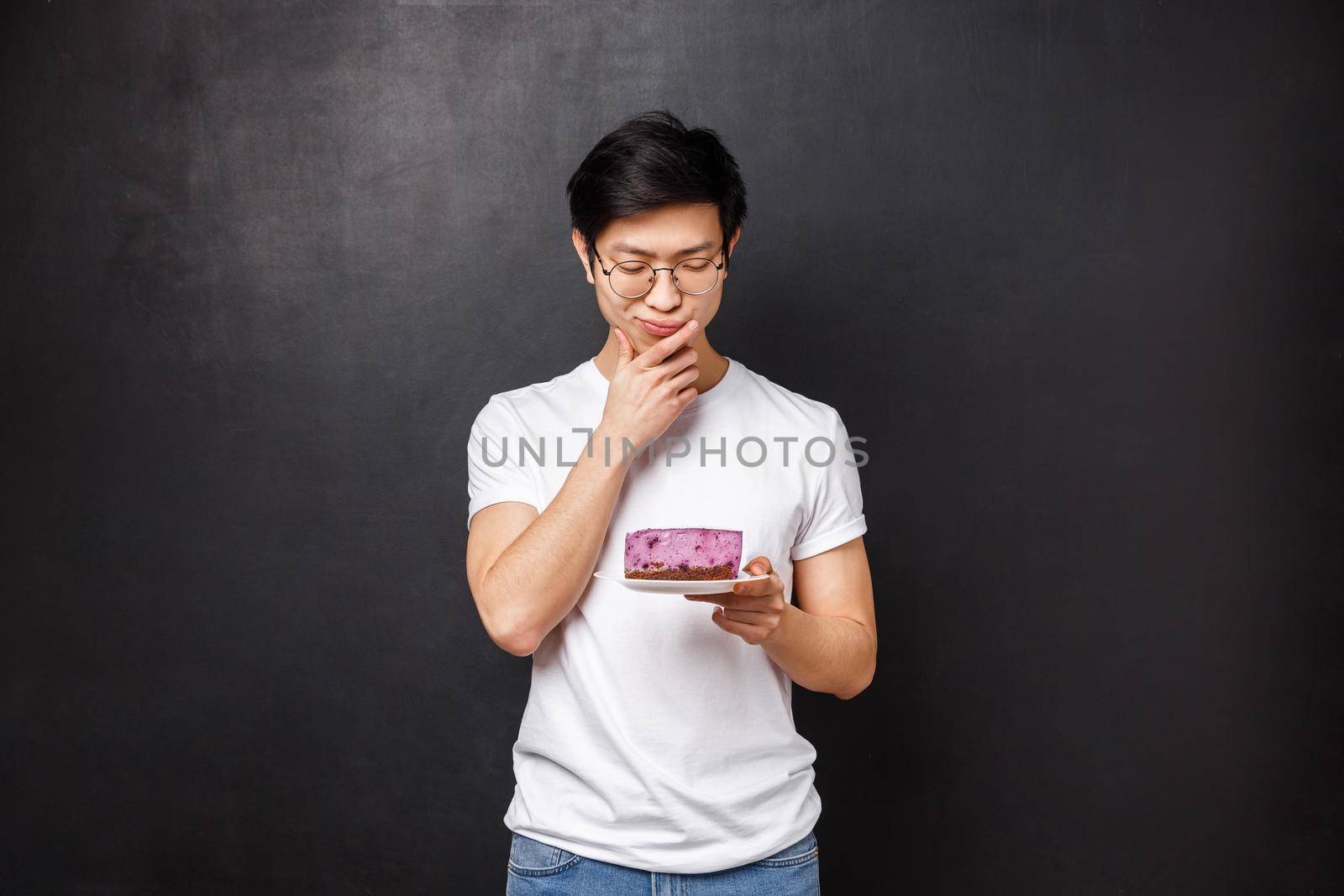Portrait of thoughtful and doubtful asian guy looking hesitant at piece of cake, thinking eat or leave it in the fridge, standing over black background, being on diet, allergic on ingridients by Benzoix