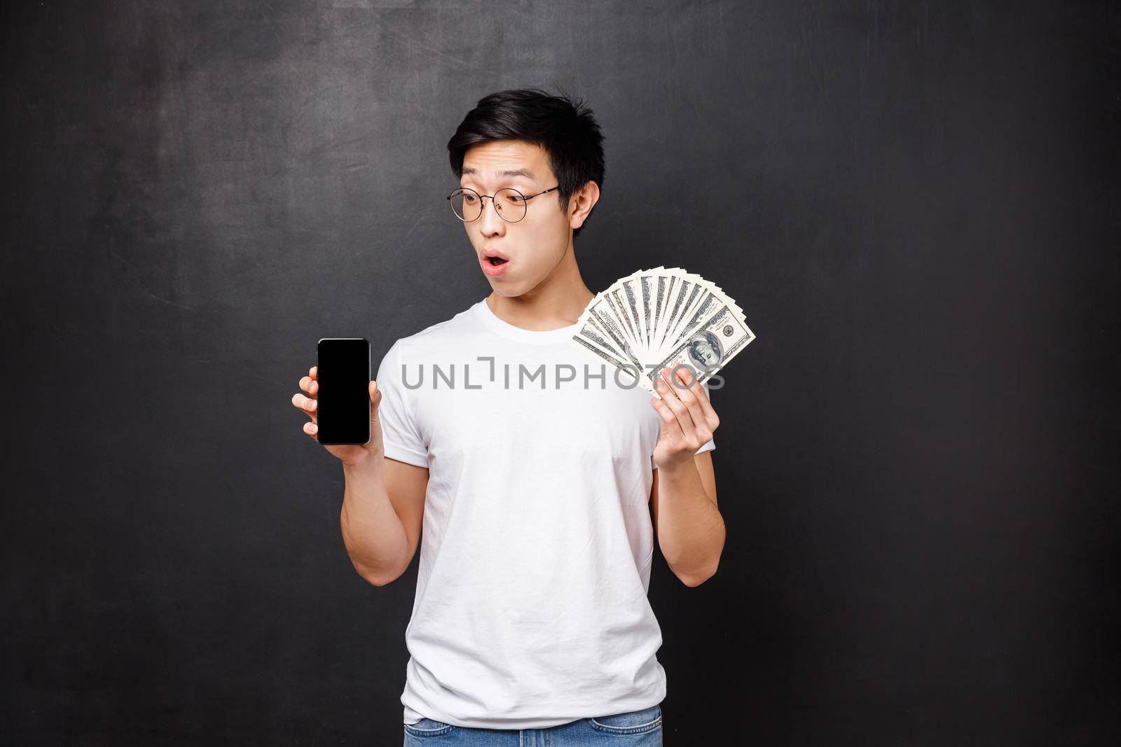 Technology, money and prizes concept. Amazed and surprised young asian guy still cant believe he won money via giveaway in internet, showing mobile phone display and dollars cash.