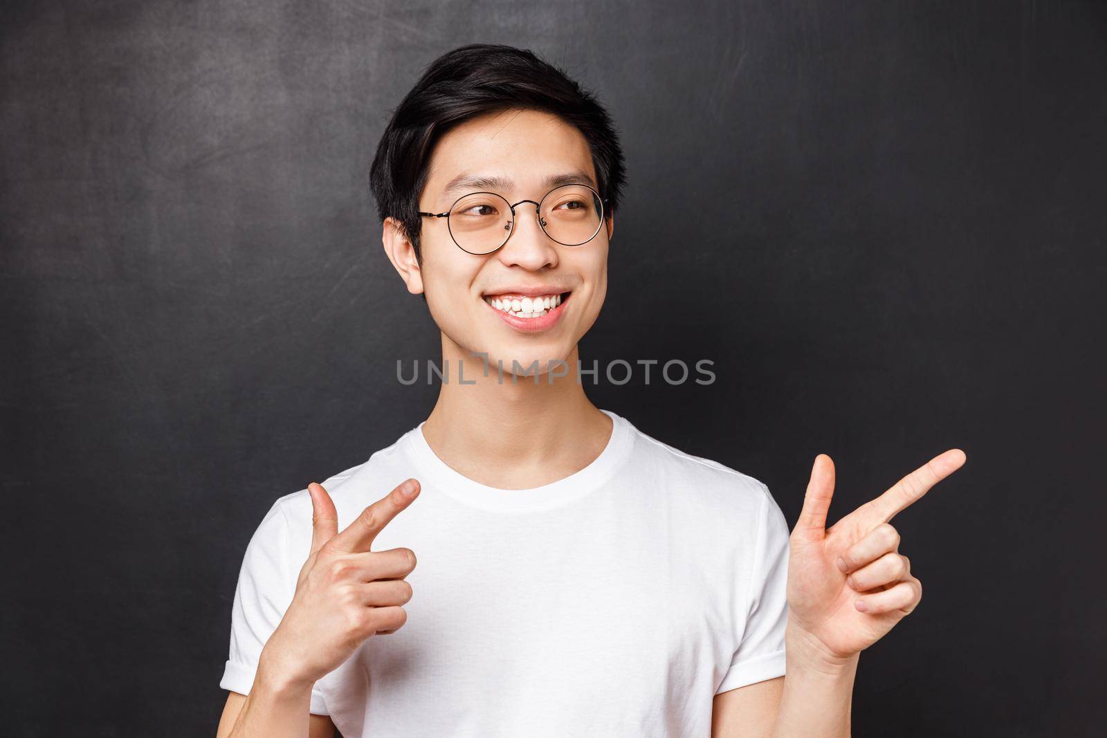 Close-up portrait of good-looking asian man in white t-shirt and glasses, spot friend on other side of room, pointing finger pistols at right side, smiling saying hi, informal greeting concept.