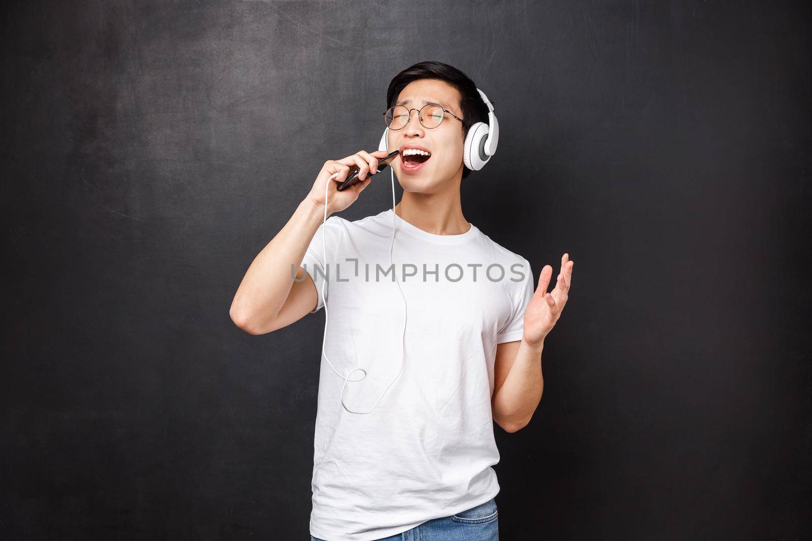 Technology, gadgets and people concept. Carefree passionate asian man listening favorite song in headphones, using mobile phone as microphone for karaoke, singing along. Copy space