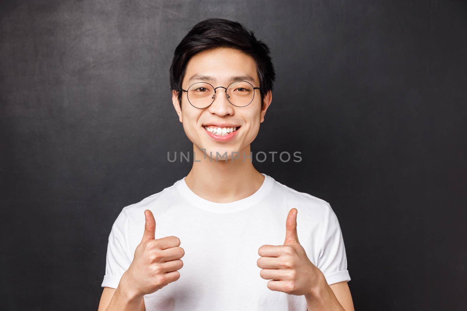 Close-up portrait of satisfied, confident asian young man in white t-shirt, show thumbs-up in approval, smiling guarantee good product, recommend service or company, black background.