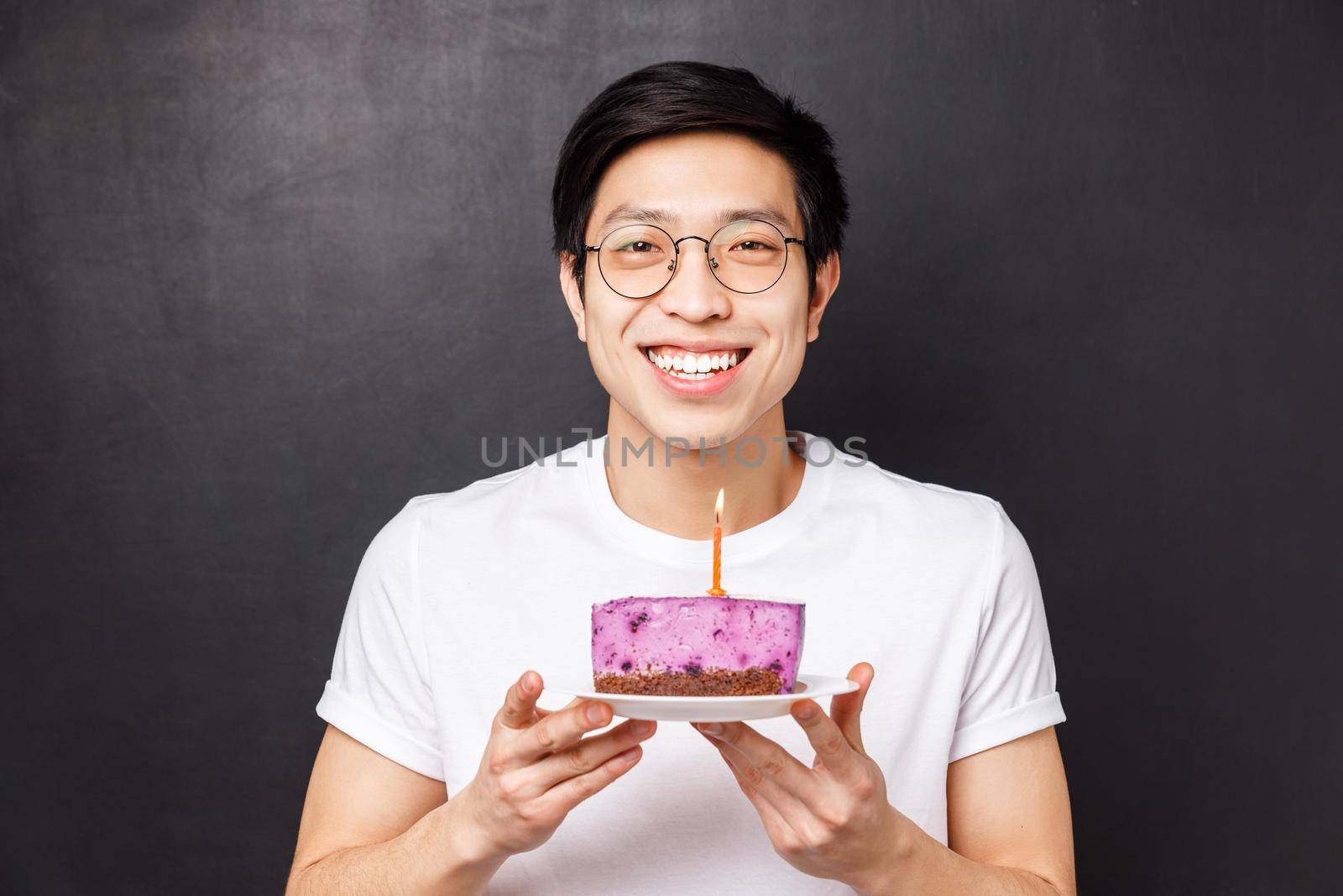 Celebration, holiday and birthday concept. Close-up portrait of happy, cheerful b-day guy holding cake with lit candle, smiling upbeat, thinking what wish make and blow-out, black background.