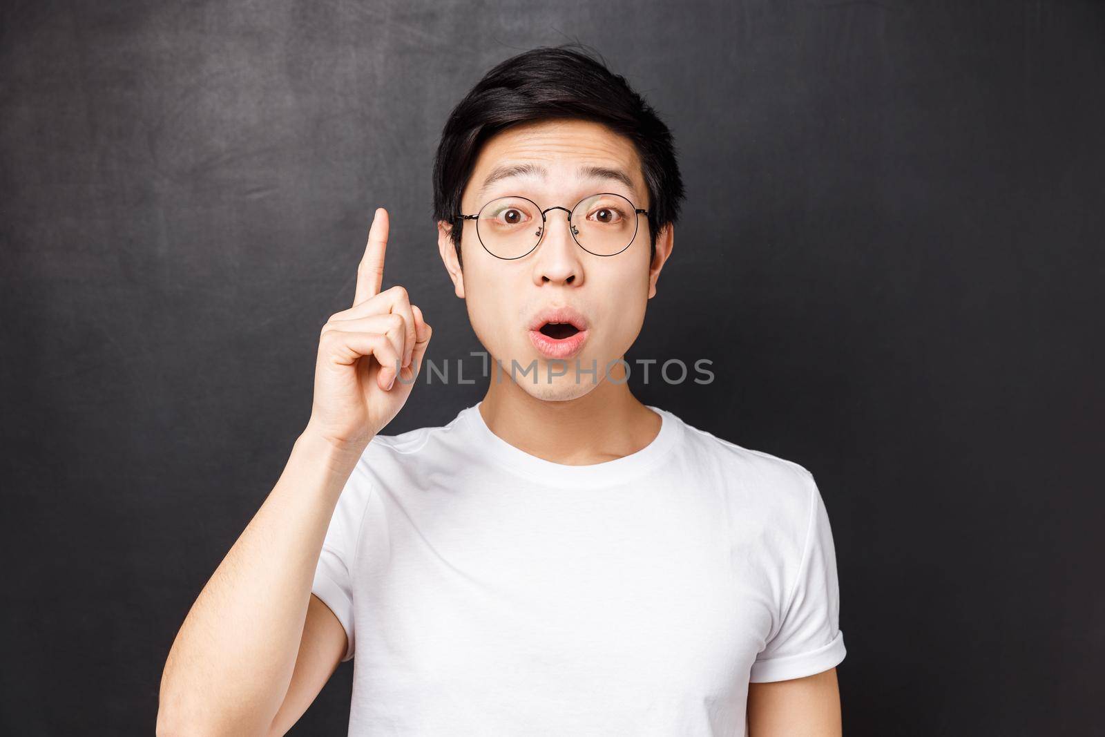 Close-up portrait of smart and creative asian man in glasses, raising index finger add suggestion, saying his opinion or thought, want to ask question, invented good plan, black background.