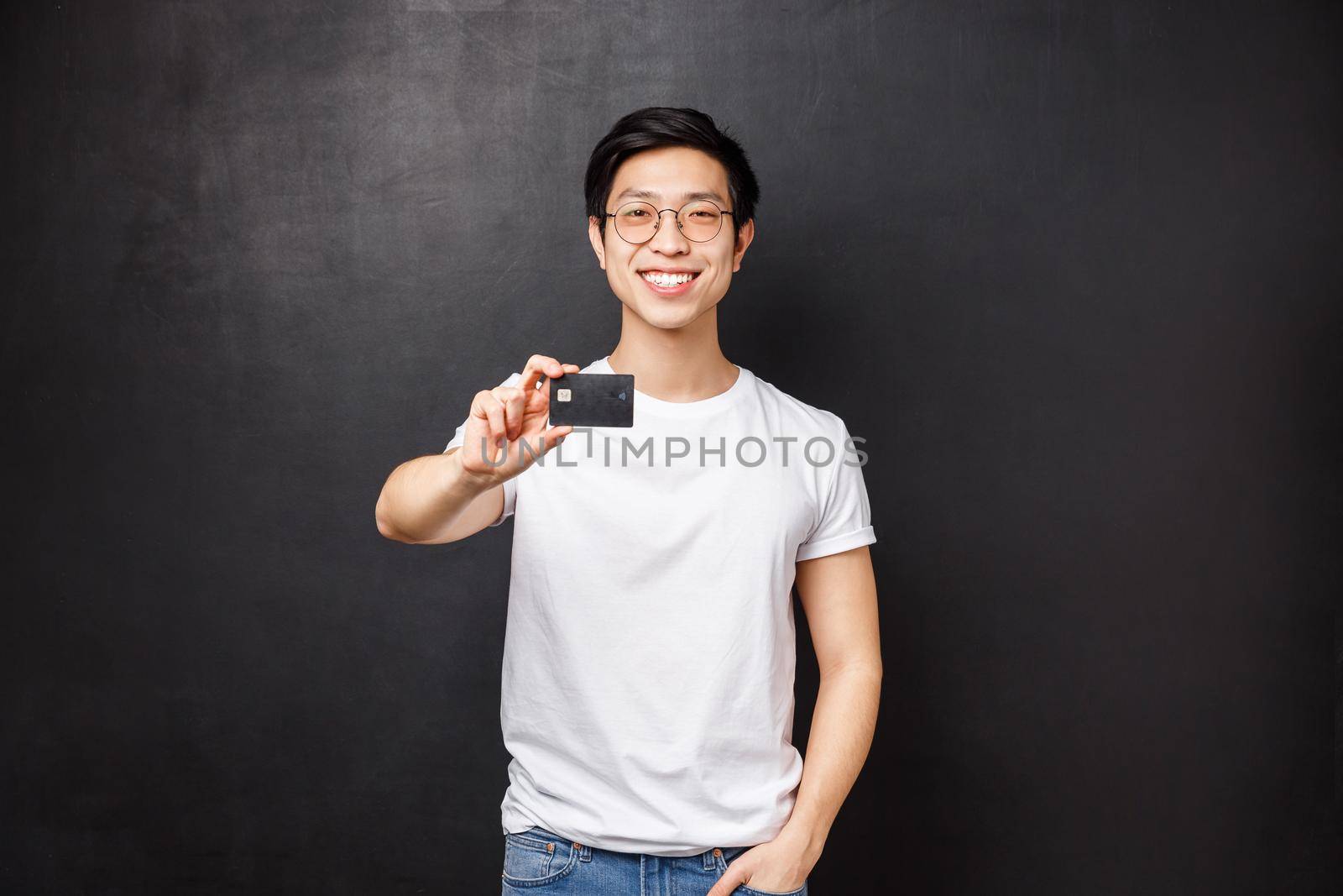 Bank, finance and payment concept. Portrait of friendly confident young asian male showing credit card at camera with pleased beaming smile, advice open account at put money, black background.