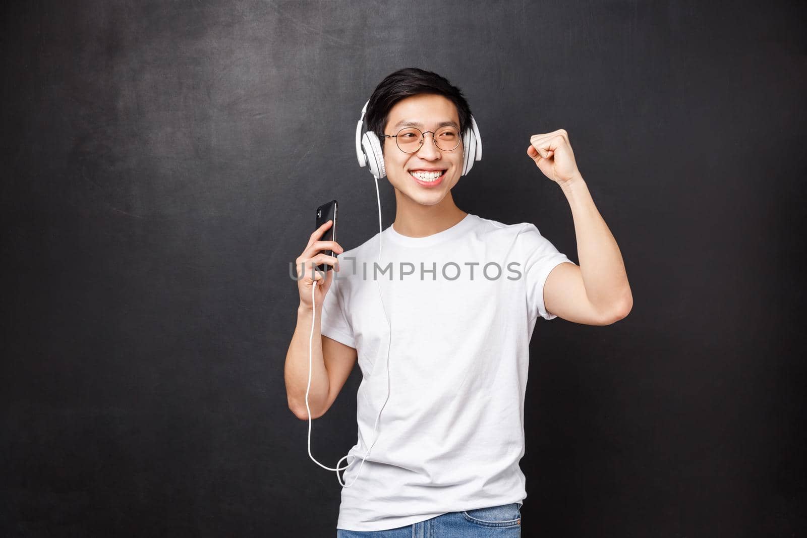 Technology, gadgets and people concept. Carefree joyful asian guy having fun, dancing to favorite single, listen music in headphones, hold mobile phone, wear earphones and smile.