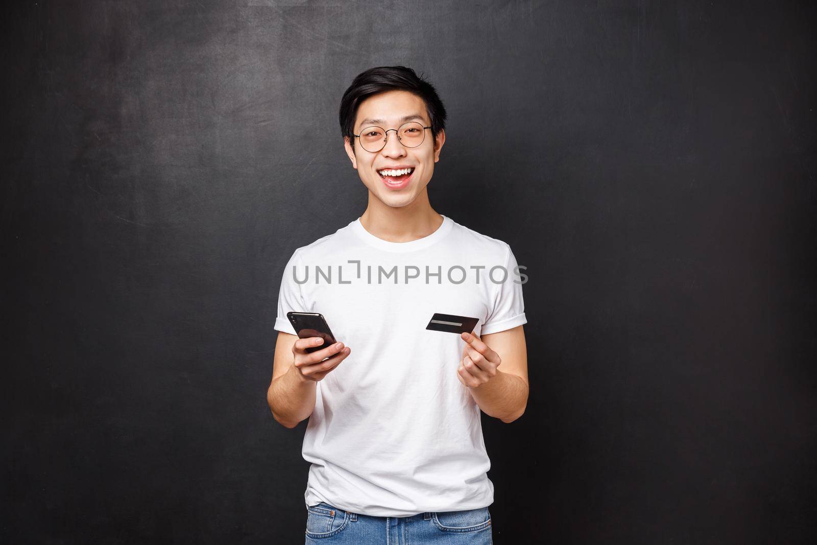 Bank, finance and payment concept. Portrait of amused handsome young asian guy holding credit card and mobile phone, smiling at camera, like shopping online, open new account, black background by Benzoix