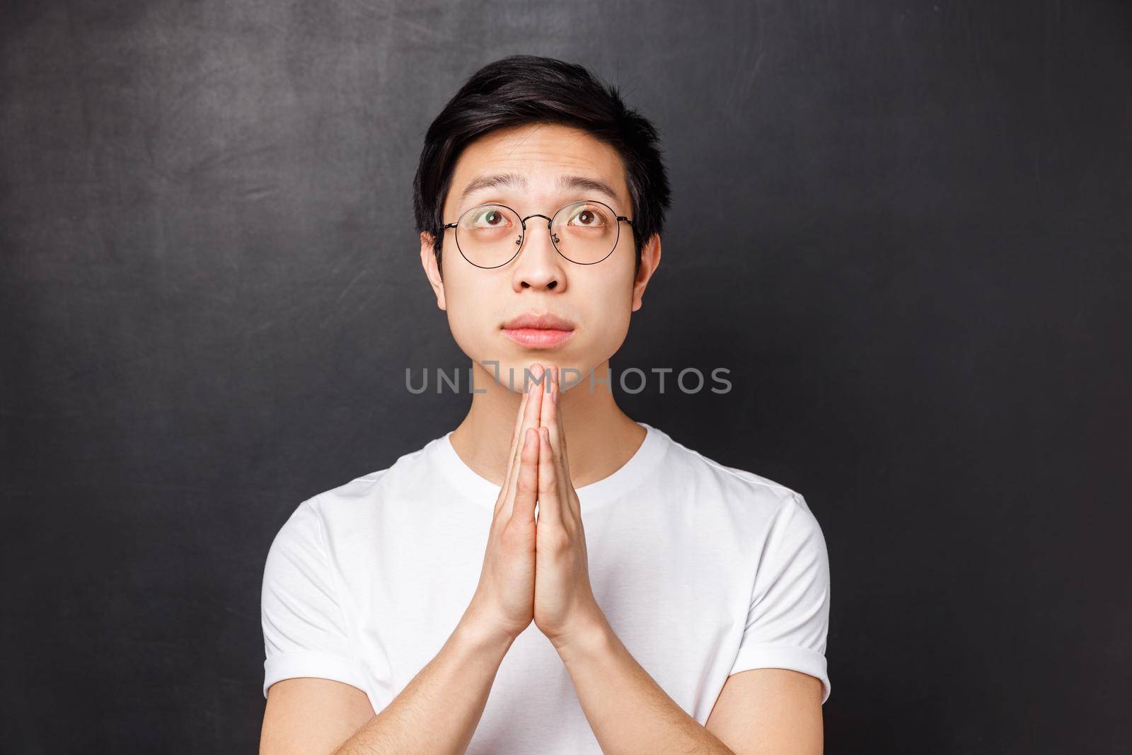 Close-up portrait of hopeful dreamy young asian guy in glasses and white t-shirt, hold hands together in pray, looking up pleading, anticipating miracle, awaiting results with hope.