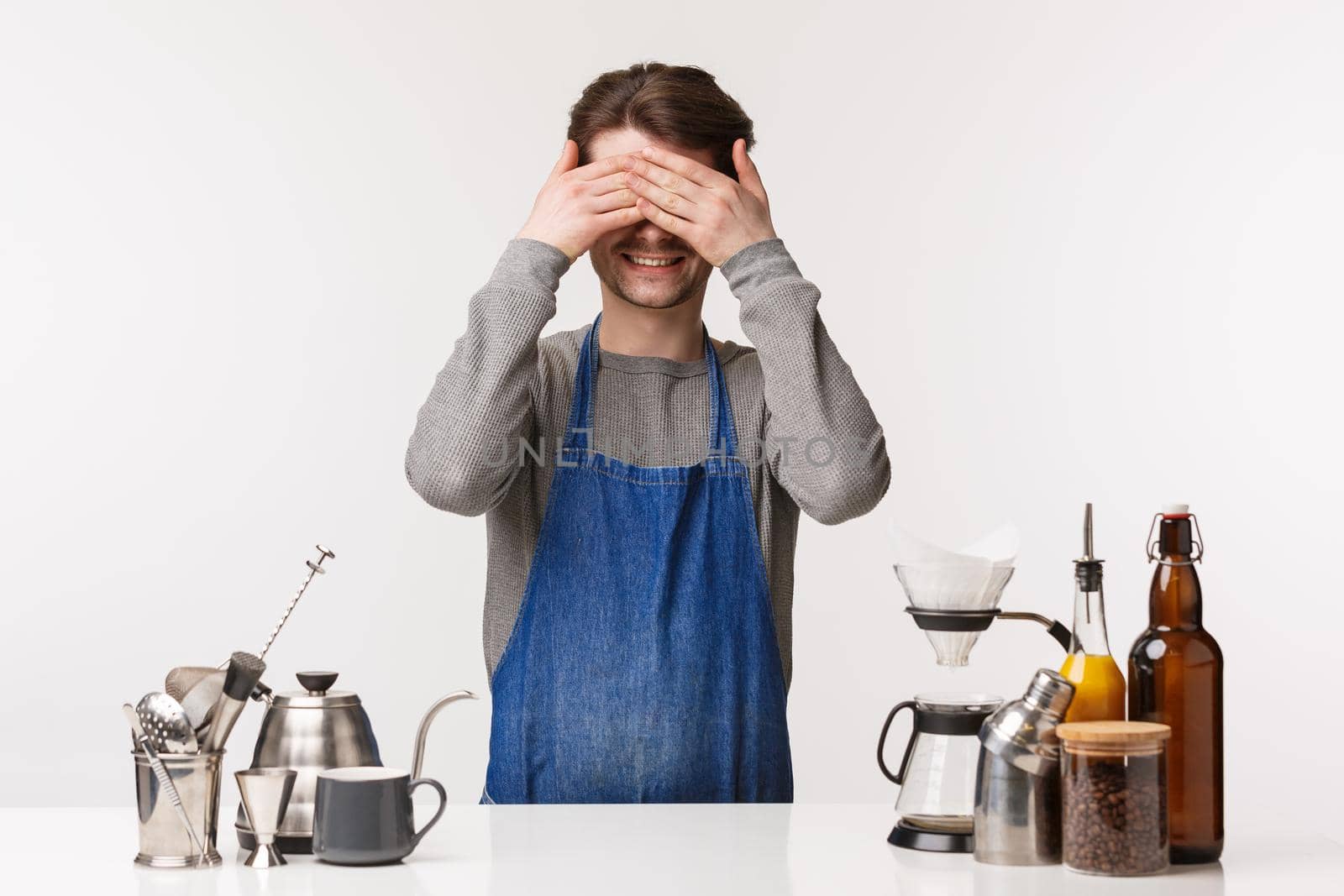 Barista, cafe worker and bartender concept. Funny and cute young male employee having fun during break, standing apron near bar counter with teapots, making coffee, close eyes and smiling.