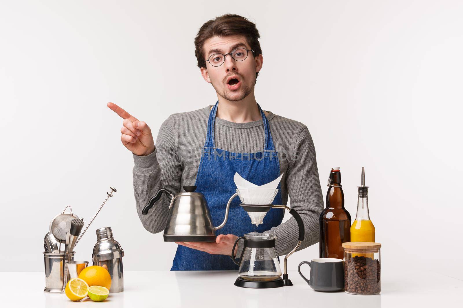 Barista, cafe worker and bartender concept. Portrait of skeptical young man pointing finger left while holding kettle, making filter coffee in chemex, standing bar counter, white background.