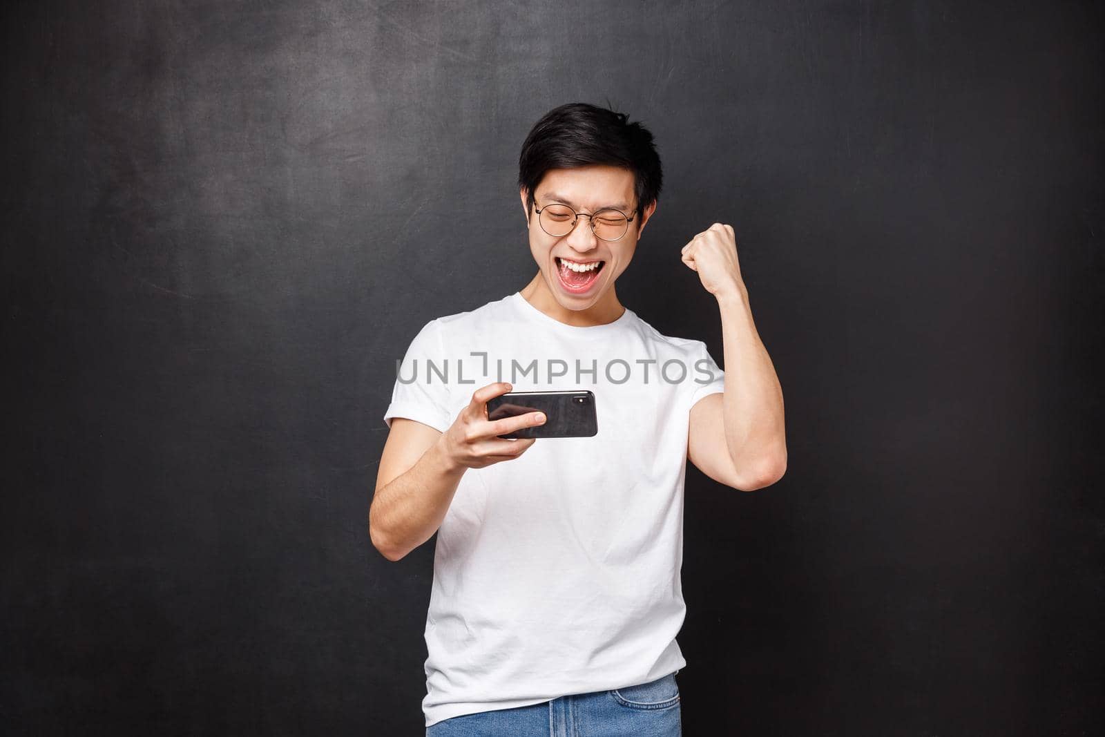 Technology, gadgets and people concept. Happy joyful lucky asian guy fist pump in celebration, hold mobile phone and smiling say yes, triumphing passed game or level, black background.