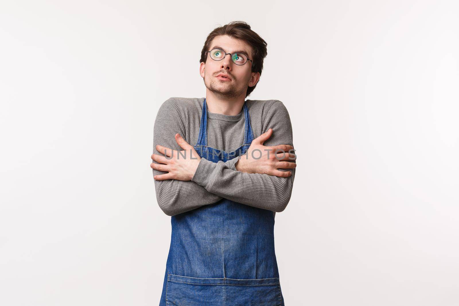 Portrait of young male student working part-time at coffee shop, have break stand outdoors in apron and look up as feel chilly, trembling and hug himself from cold weather, white background.