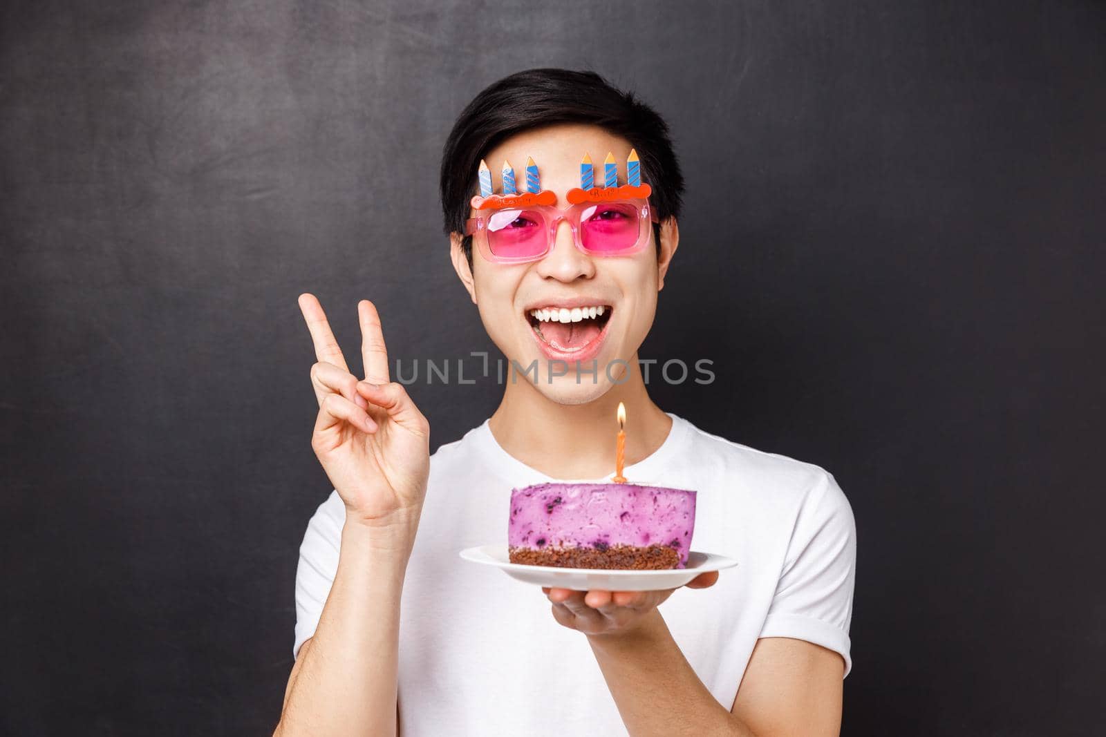 Celebration, holiday and birthday concept. Close-up portrait of joyful funny asian man having fun, enjoying b-day party, show peace sign smiling and hold cake with lit candle, make wish.