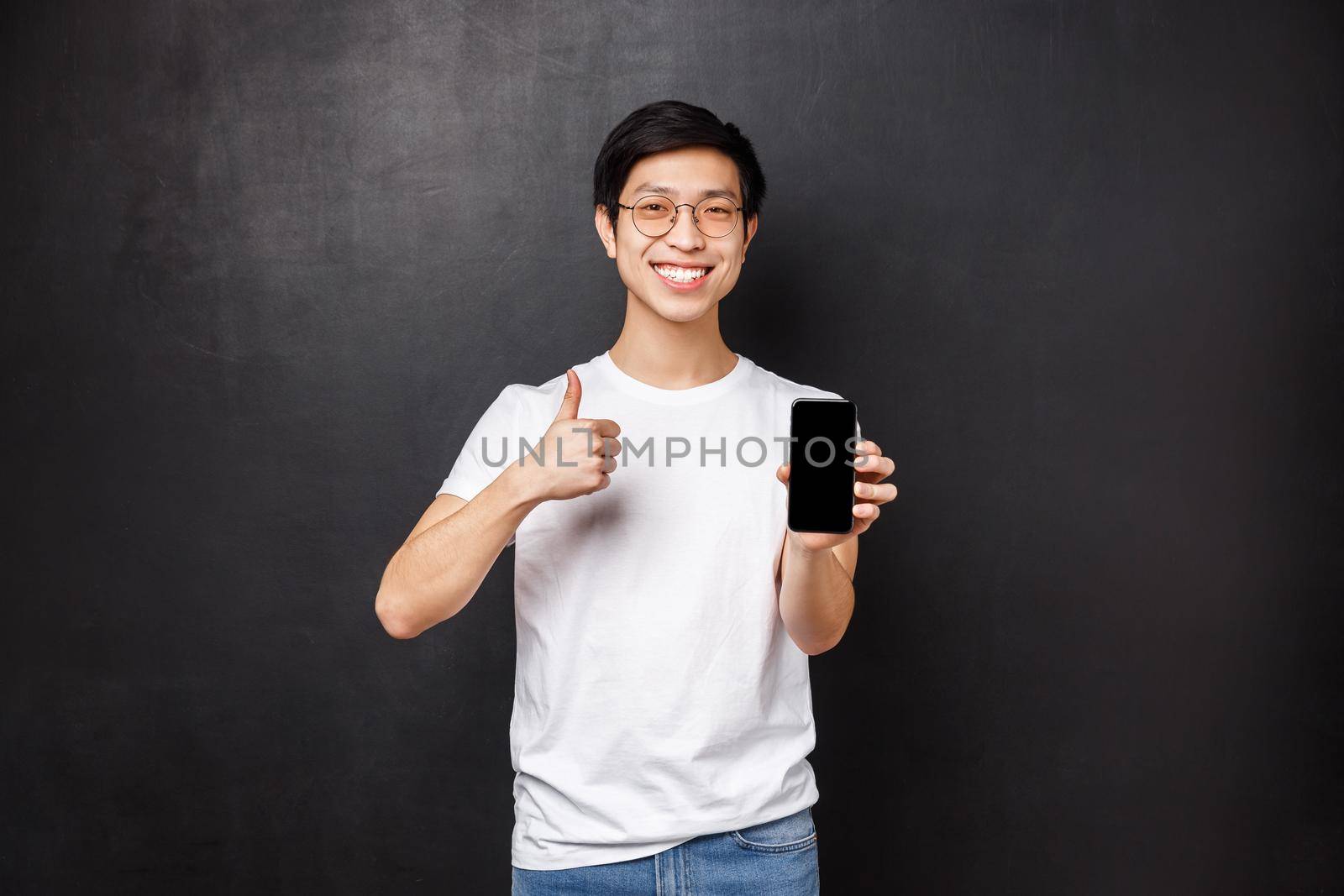 Technology, gadgets and people concept. Assertive cute asian guy in t-shirt and glasses, recommend online store or application for mobile phone, show thumbs-up and smartphone display.