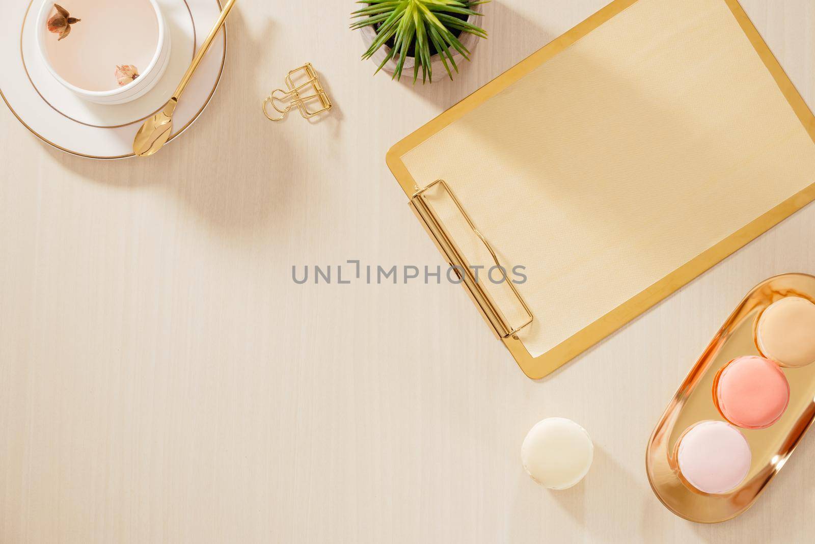 Modern gold stylized home office desk with folder, macaroons, coffee mug on beige background. Flat lay, top view lifestyle concept.`