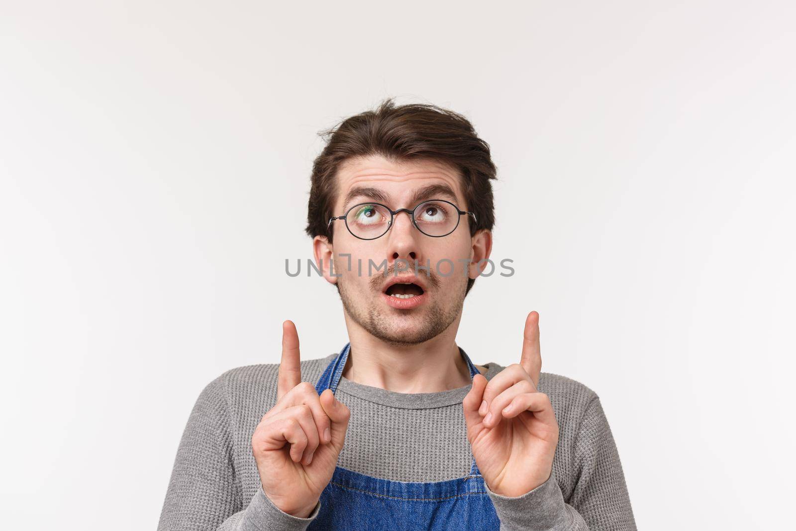 What is it. Intrigued and excited young surprised man in glasses and apron looking at something strange and curious, pointing fingers up, standing white background uncertain and careful.