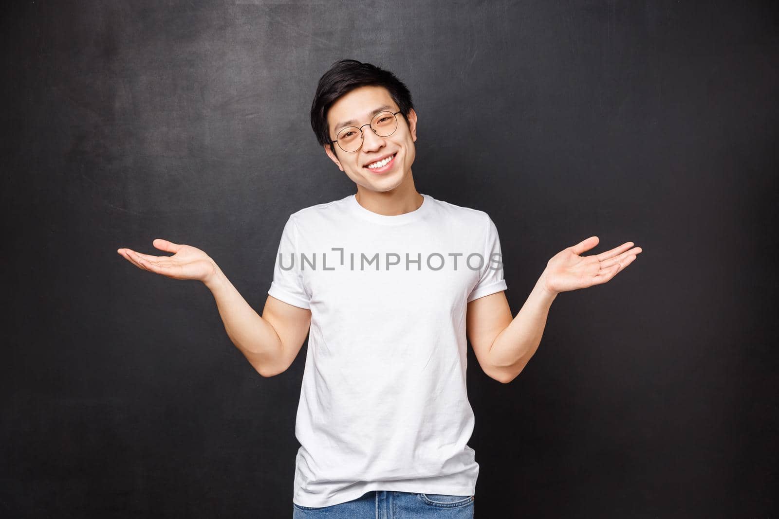No big dea. Handsome carefree and unbothered asian guy shrugging and spread hands sideways clueless, have no idea, dont know anything and not care at all, black background, smiling.