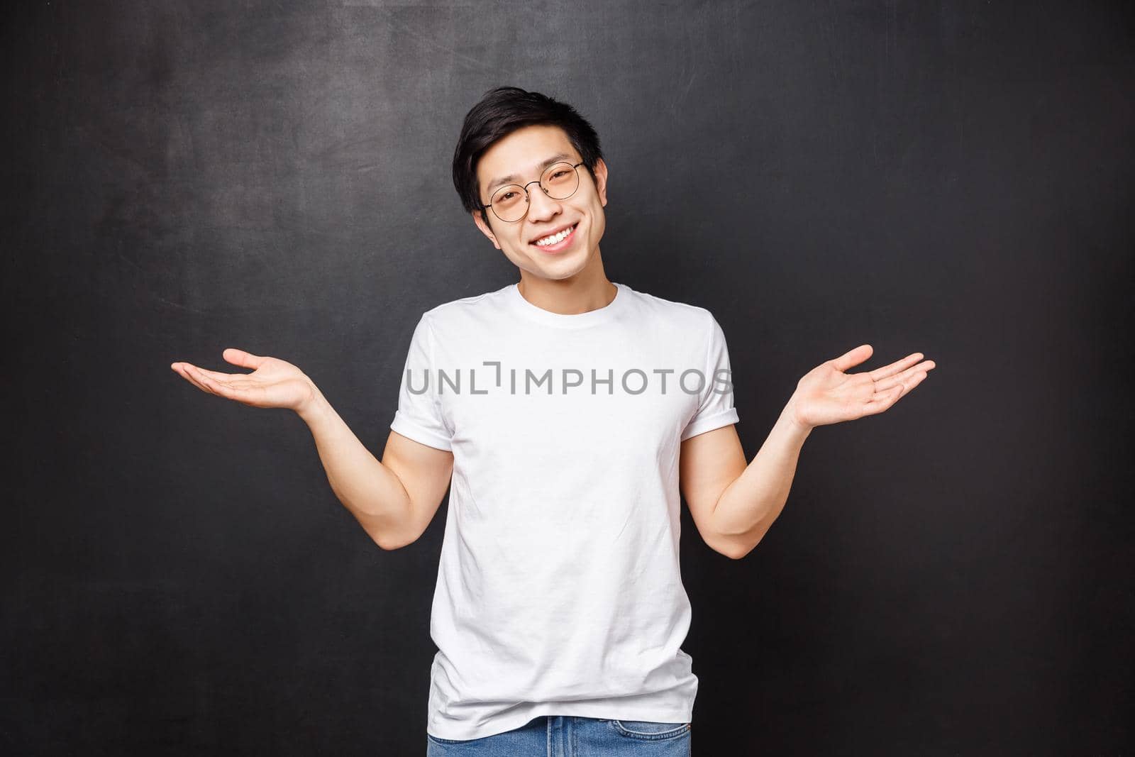 No big dea. Handsome carefree and unbothered asian guy shrugging and spread hands sideways clueless, have no idea, dont know anything and not care at all, black background, smiling.