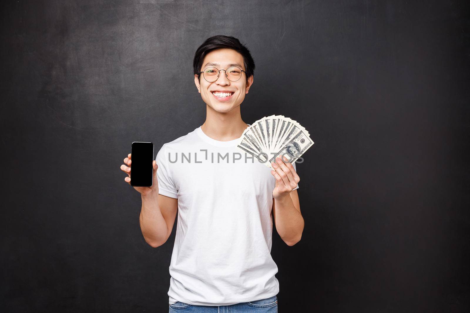 Portrait of proud rich young man showing dollars, fan of money and mobile phone display, smiling as bragging where he won receive prize cash, standing satisfied over black background.