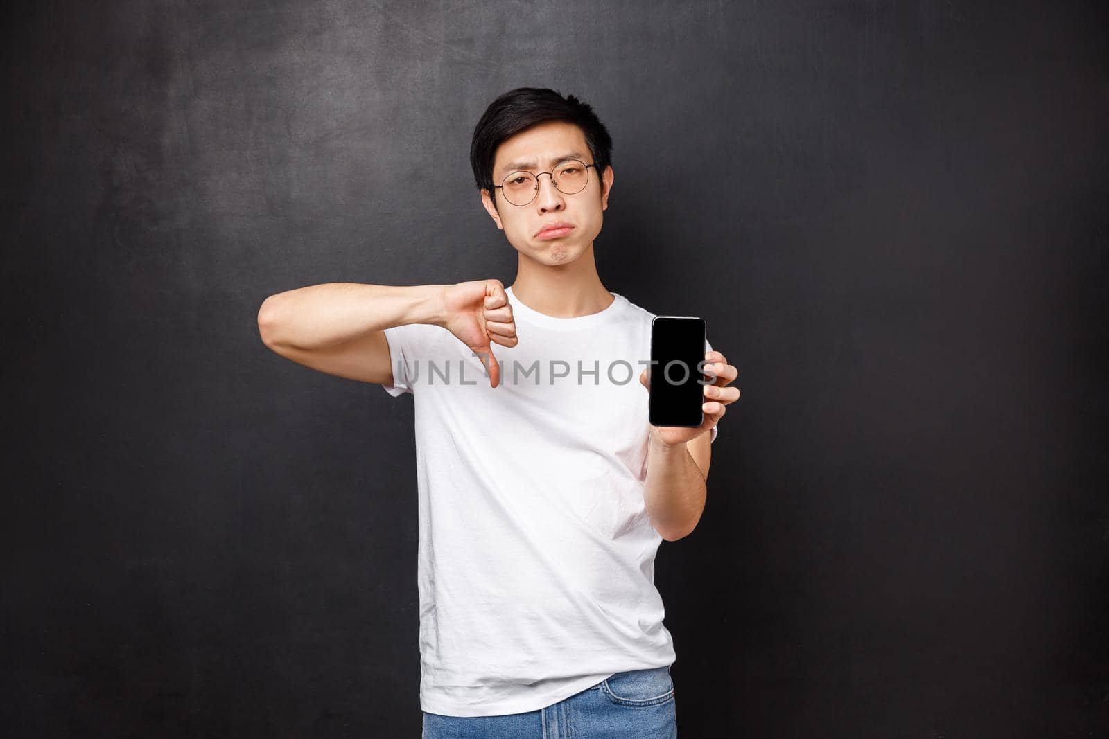 Technology, gadgets and people concept. Gloomy dissatisfied asian guy rate bad application, being disappointed with service awful quality, show mobile phone screen and thumbs-down, black background by Benzoix