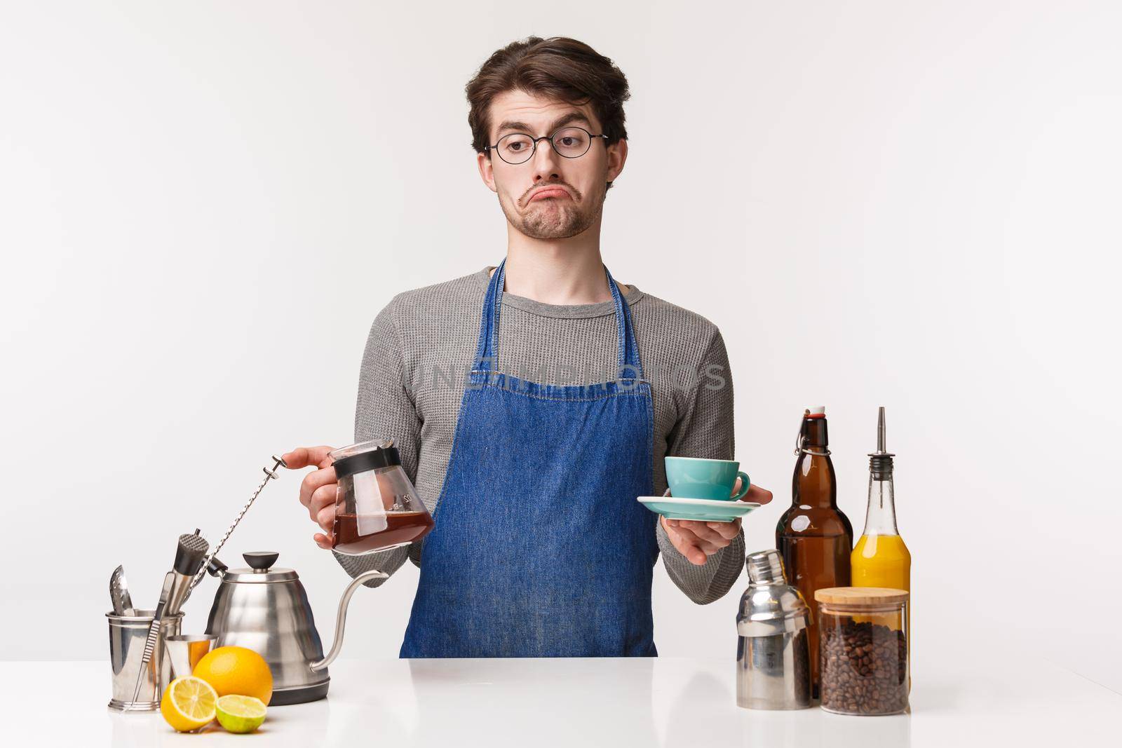 Barista, cafe worker and bartender concept. Portrait of indecisive careless young bored male employee grimacing look away uncertain, hold kettle with filter coffee and cup, white background.