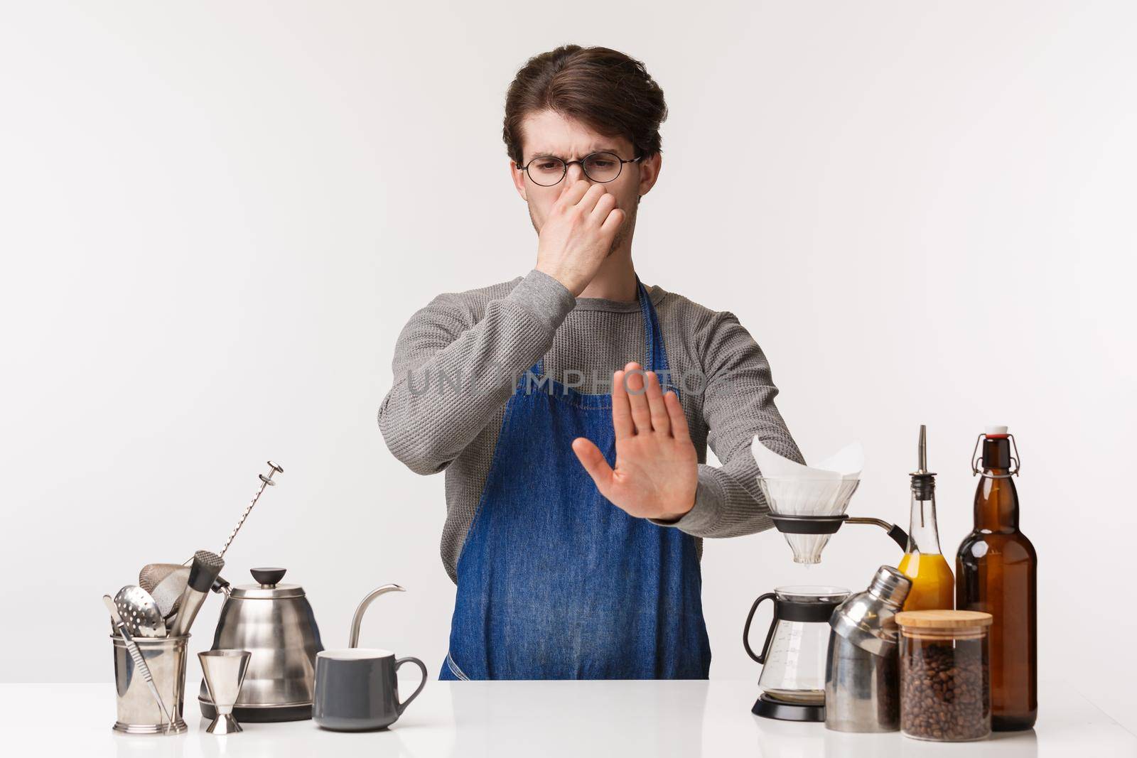 Barista, cafe worker and bartender concept. Portrait of displeased and reluctant young male in apron, standing near bar counter with goods, coffee hear nasy awful smell, show stop sign.